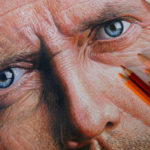 Detail realistické kresby mužské tváře se zaměřením na modré oči a vrásky. Na pravé straně díla jsou umístěny barevné tužky různých odstínů.