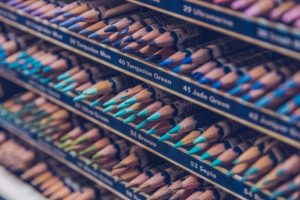 Detailní záběr na řady barevných tužek uspořádaných podle odstínů, s nápisy na policích, které označují barvy jako "tyrkysově zelená", "nefritově zelená", "bronzová" a "sépiová". Hroty tužek směřují ven.