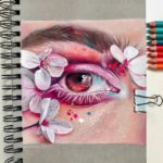 Realistická kresba lidského oka zdobeného růžovými očními stíny a obklopeného bílými okvětními lístky třešňových květů, vytvořená na stránce spirálového skicáku s barevnými tužkami vedle.