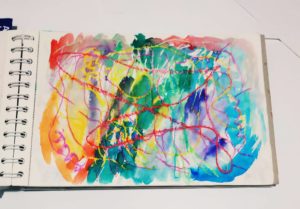 V otevřeném skicáku je zobrazena barevná abstraktní malba s vířícími červenými liniemi na bílém papíře v zářivých odstínech modré, zelené, žluté, oranžové a růžové akvarelové barvy.