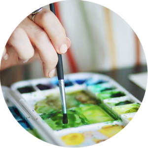 Ruka držící štětec se namáčí do zelené akvarelové barvy na malířské paletě, na které jsou vidět různé odstíny zelené, žluté a modré.