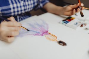 Osoba v modré kostkované košili maluje vodovými barvami a tužkou postavu v šatech na bílý papír. Pozornost je zaměřena na její ruce a umělecké dílo, přičemž barvy jsou viditelné vedle papíru.