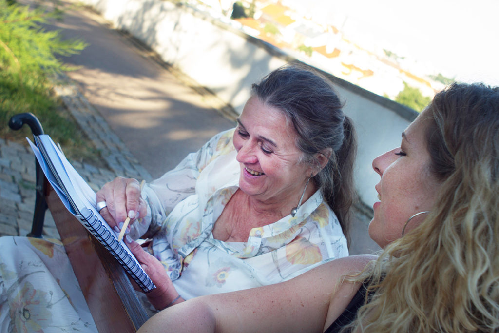 Dvě ženy sedí na lavičce venku, usmívají se a společně si prohlížejí spirálový zápisník. Jedna z žen ukazuje na zápisník a zdá se, že si užívají příjemný rozhovor.