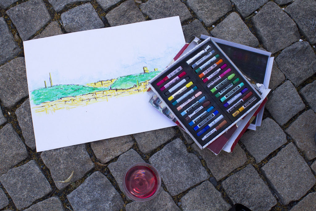 Na šedé dlažbě je položena kresba zelené krajiny na bílém papíře, otevřená krabice s barevnými pastely a sklenice s růžovou tekutinou.