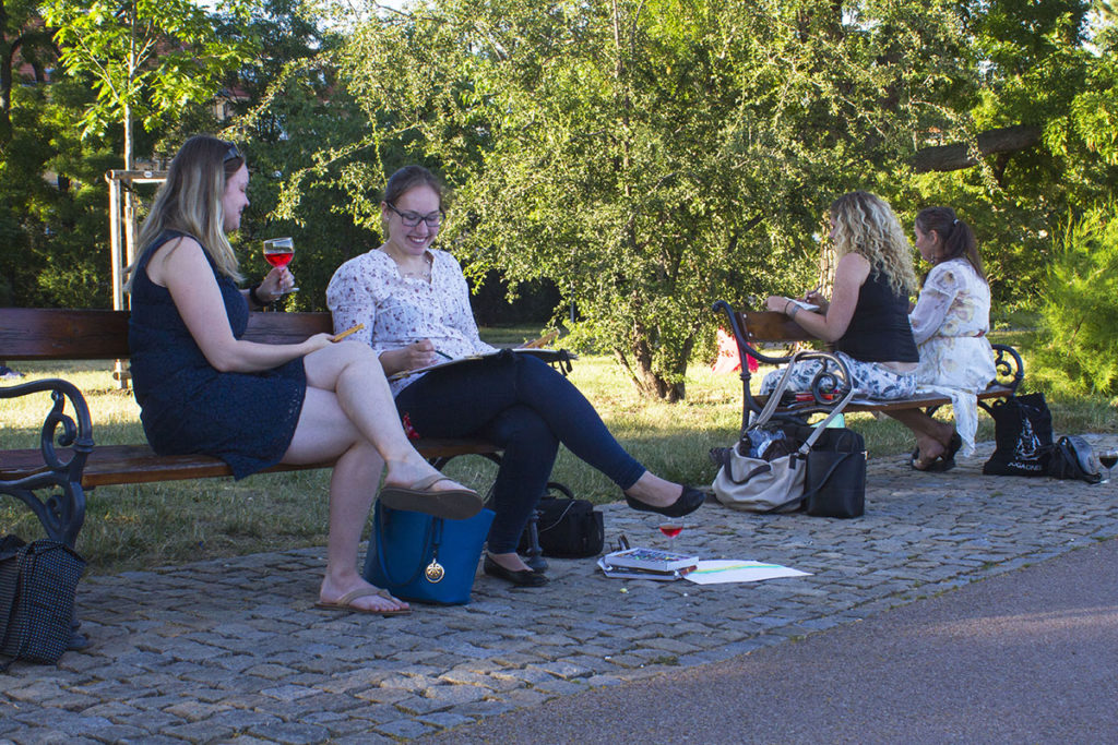 Čtyři ženy sedí za slunečného dne na lavičkách v parku. Dvě ženy si povídají a drží nápoje, jedna píše do notebooku a druhá píše na notebooku. Na zemi poblíž jsou rozházené tašky a papíry.