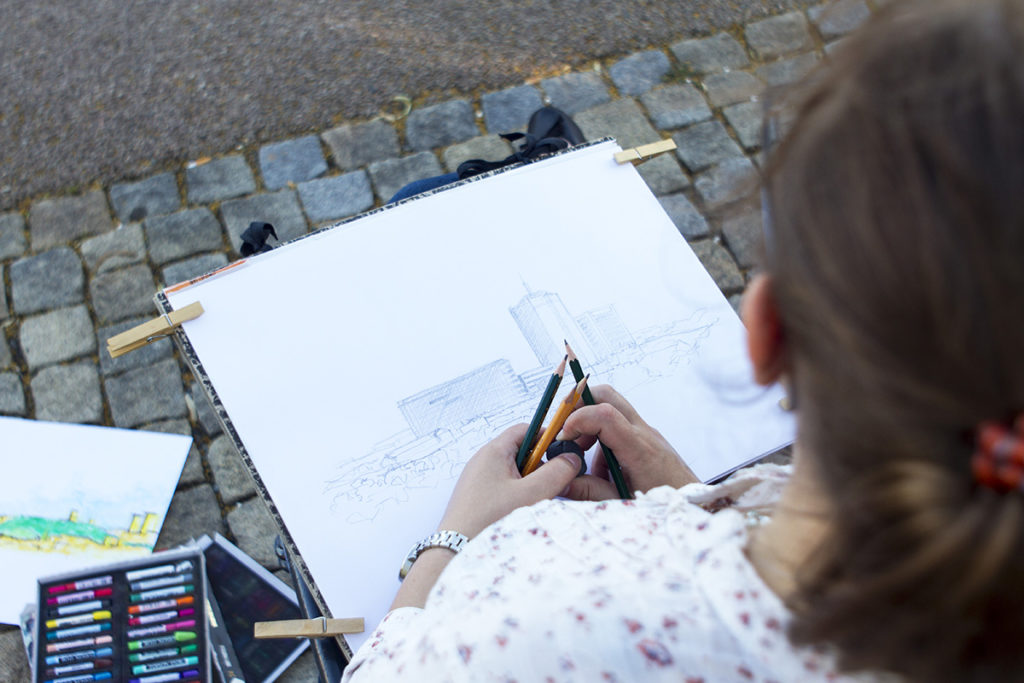 Osoba s hnědými vlasy svázanými dozadu kreslí venku na velký list papíru budovy, sedí na dlažebních kostkách. Vedle nich leží výtvarné potřeby včetně barevných tužek a náčrtníku. Scéna působí klidně a tvůrčím dojmem.