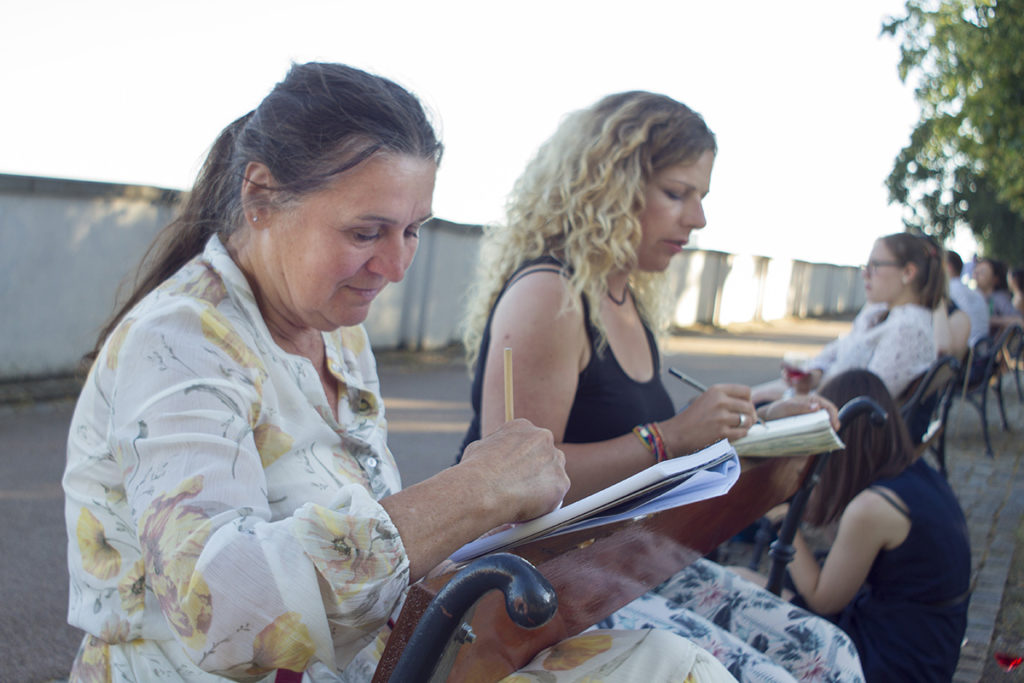 Dvě ženy sedí na lavičce venku a soustředěně si kreslí nebo píší do sešitů. V pozadí sedí další lidé a scénu osvětluje sluneční světlo.