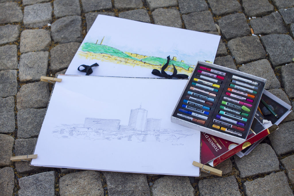 Dva skicáky s krajinnými kresbami leží na dlažbě; v jednom je kolorovaná venkovská scéna, ve druhém tužkou kreslená městská krajina. Vedle leží otevřená krabice s barevnými olejovými pastely.