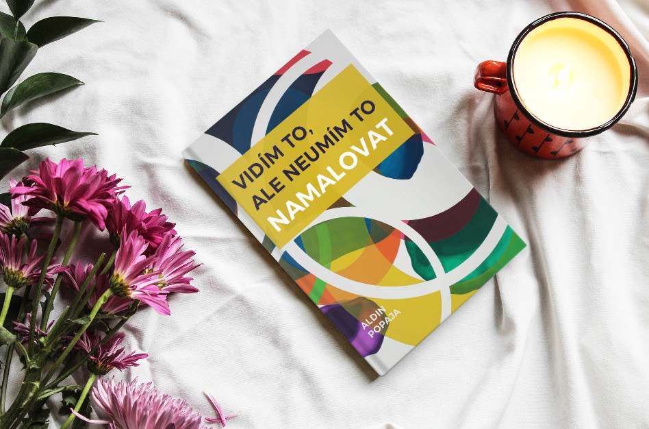 Na bílé látce leží kniha s barevným abstraktním obalem, vedle ní fialové květy, oranžová svíčka v hrnku a zelené listy. Název knihy je v češtině: "Vidím to, ale neumím to namalovat.