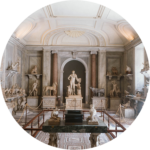 Ve velkolepé muzejní místnosti jsou vystaveny klasické mramorové sochy včetně centrální stojící postavy, zvířat a různých soch na policích a podstavcích, obklopené zdobnou architekturou a měkkým osvětlením.