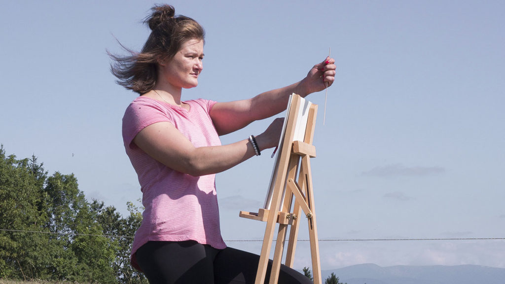 Žena v růžové košili stojí venku a upravuje prázdné plátno na dřevěném stojanu. V pozadí jsou vidět stromy a modrá obloha s lehkými mraky.