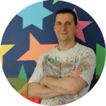 Před barevným pozadím s velkými hvězdami a geometrickými obrazci stojí osoba s dlouhými tmavými vlasy a zkříženýma rukama, která má na sobě košili potřísněnou barvou.