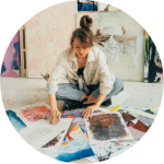 Žena sedí se zkříženýma nohama na podlaze a probírá se různými barevnými obrazy a kresbami, za ní jsou vystavena další umělecká díla ve světlém ateliéru.