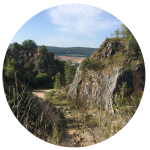 Kruhová fotografie zobrazující skalnatou krajinu se strmými útesy, suchou trávou a řídkými stromy pod modrou oblohou s roztroušenými mraky; středem se vine polní cesta.