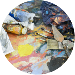 Nepořádek na pracovišti umělce s tubami s barvou, štětci, paletovým nožem a hadříkem na povrchu pokrytém barevnými cákanci barvy a žlutou skvrnou od barvy.