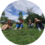 Čtyři mladí lidé sedí venku na trávě a kreslí na velkou tabuli. V pozadí jsou stromy a částečně zatažená obloha, což naznačuje prostředí parku. Nálada působí uvolněně a soustředěně.