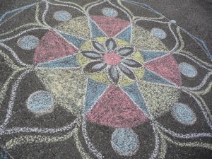 Barevný křídový design rangoli s kruhovými a květinovými vzory v růžové, žluté, modré a bílé barvě, nakreslený na asfaltu. Symetrické umělecké dílo obsahuje okvětní lístky a geometrické tvary vyzařující ze středu.