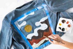 Osoba maluje štětcem a paletou barev na záda džínové bundy barevnou krajinu se žlutým sluncem, bílými mraky a červenohnědými kopci. Modrá malířská páska rámuje namalovanou plochu.