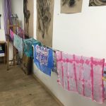 V uměleckém ateliéru visí na šňůře několik barevných, ručně malovaných hedvábných šátků s různými vzory. Na stěně za nimi jsou vystaveny figurální kresby uhlím a vlevo od nich stojí stojan.