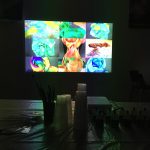 Tmavá místnost se stolem se štětci, plastovými kelímky a lahvemi v popředí. Na světlé obrazovce v pozadí se zobrazuje mřížka s živými abstraktními obrázky v pestrých barvách.