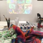 Stůl s barevnými malovanými šátky, sklenicemi se štětci, lahvemi s barvami na textil a průhlednými plastovými kelímky. Za ním se na bílé stěně promítají obrázky malovaných šátků.