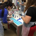 Lidé v plastových zástěrách a rukavicích malují na obdélníková plátna u stolu pokrytého výtvarnými potřebami včetně štětců, kelímků a lahví. Atmosféra vypadá jako ve skupinové výtvarné třídě nebo dílně.