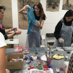 Čtyři lidé v zástěrách pracují u stolu s výtvarnými potřebami, barvami a látkami. Jedna žena drží modrou látku a usmívá se, zatímco ostatní se věnují svým projektům. Na stěně v pozadí visí kresby uhlím.