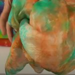 Ruka drží velkou zmačkanou látku v oranžové, zelené a tyalové barvě. V pozadí je na světlém podkladu položena další zmačkaná tkanina v duhových barvách.