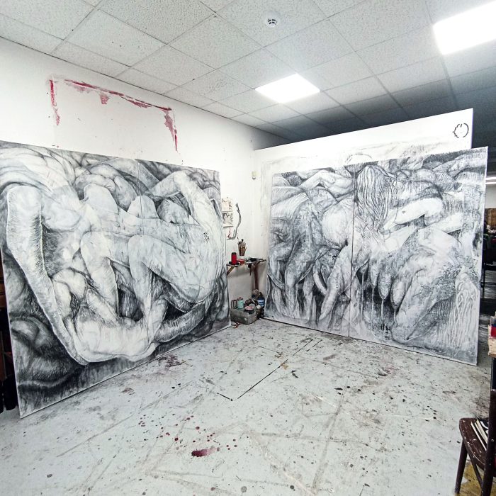 Ateliér se dvěma velkými abstraktními černobílými kresbami s propletenými organickými formami, výtvarné potřeby a židle rozeseté po podlaze potřísněné barvou v jasně osvětlené místnosti.