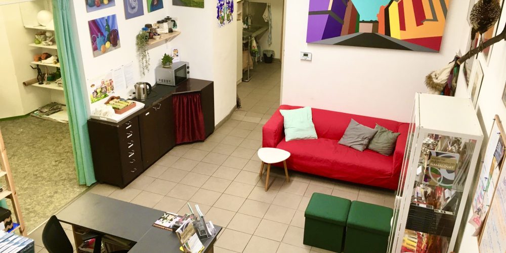 Světlý, útulný umělecký ateliér s barevnými obrazy na bílých stěnách, červenou pohovkou, zelenými otomany, malým bílým stolkem, psacím stolem, policemi s výtvarnými potřebami a kuchyňským koutem s varnou konvicí a hrnky.