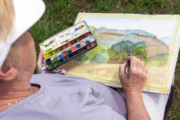 Starší člověk sedí venku a maluje vodovými barvami krajinu se stromy a kopci. V jedné ruce drží štětec a v druhé paletu s barvami, v pozadí je vidět tráva.