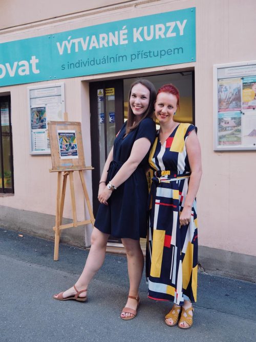 Dvě ženy se usmívají a společně pózují na chodníku před budovou s nápisem "Výtvarné kurzy". Jedna má na sobě černé šaty, druhá barevné vzorované šaty. Za nimi stojí stojan s uměleckými díly.